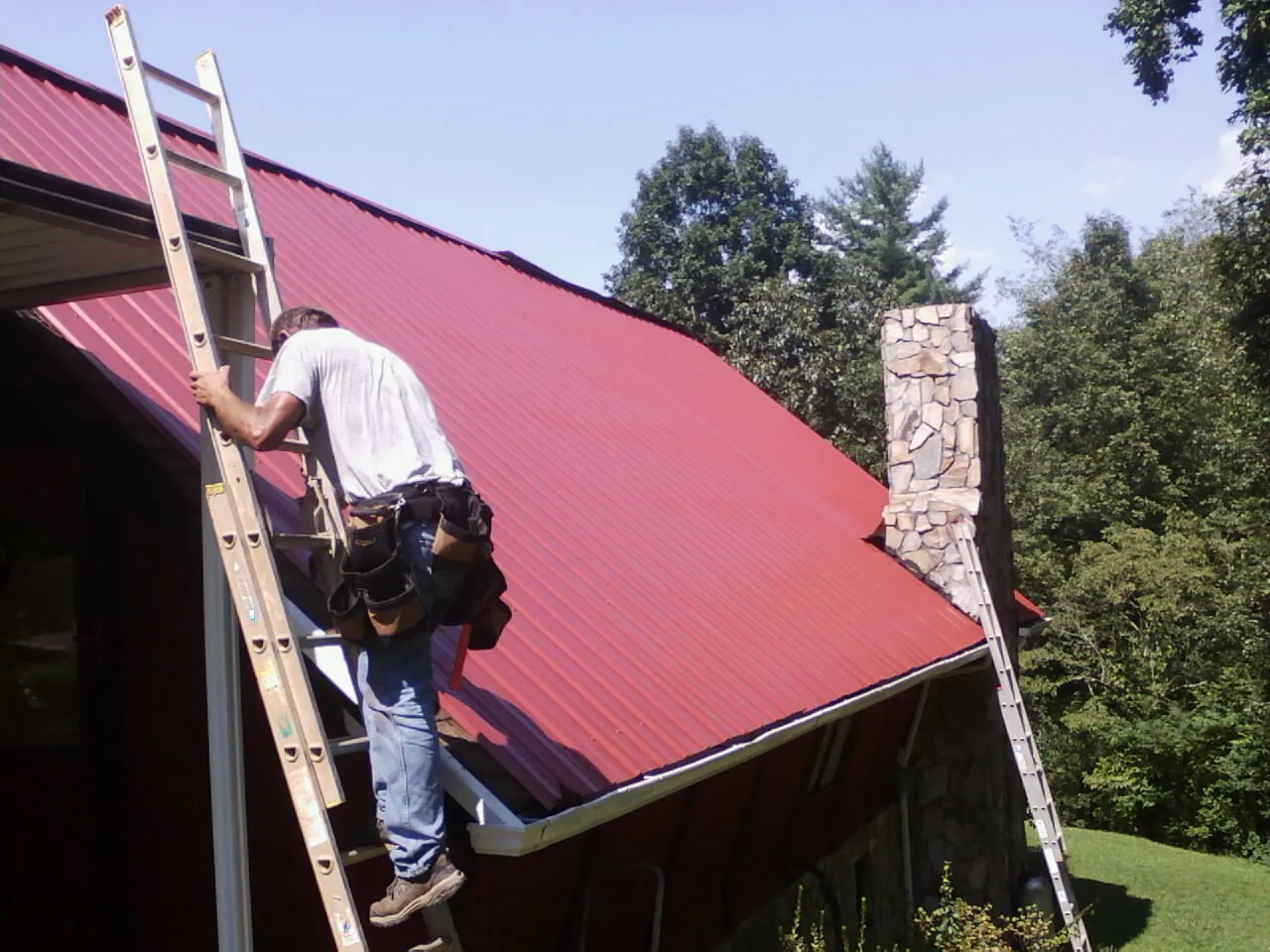 Roofer on a ladder beside a red metal roof and stone chimney — Soffit & Fascia Repair by Roofing Brothers in Silverton
